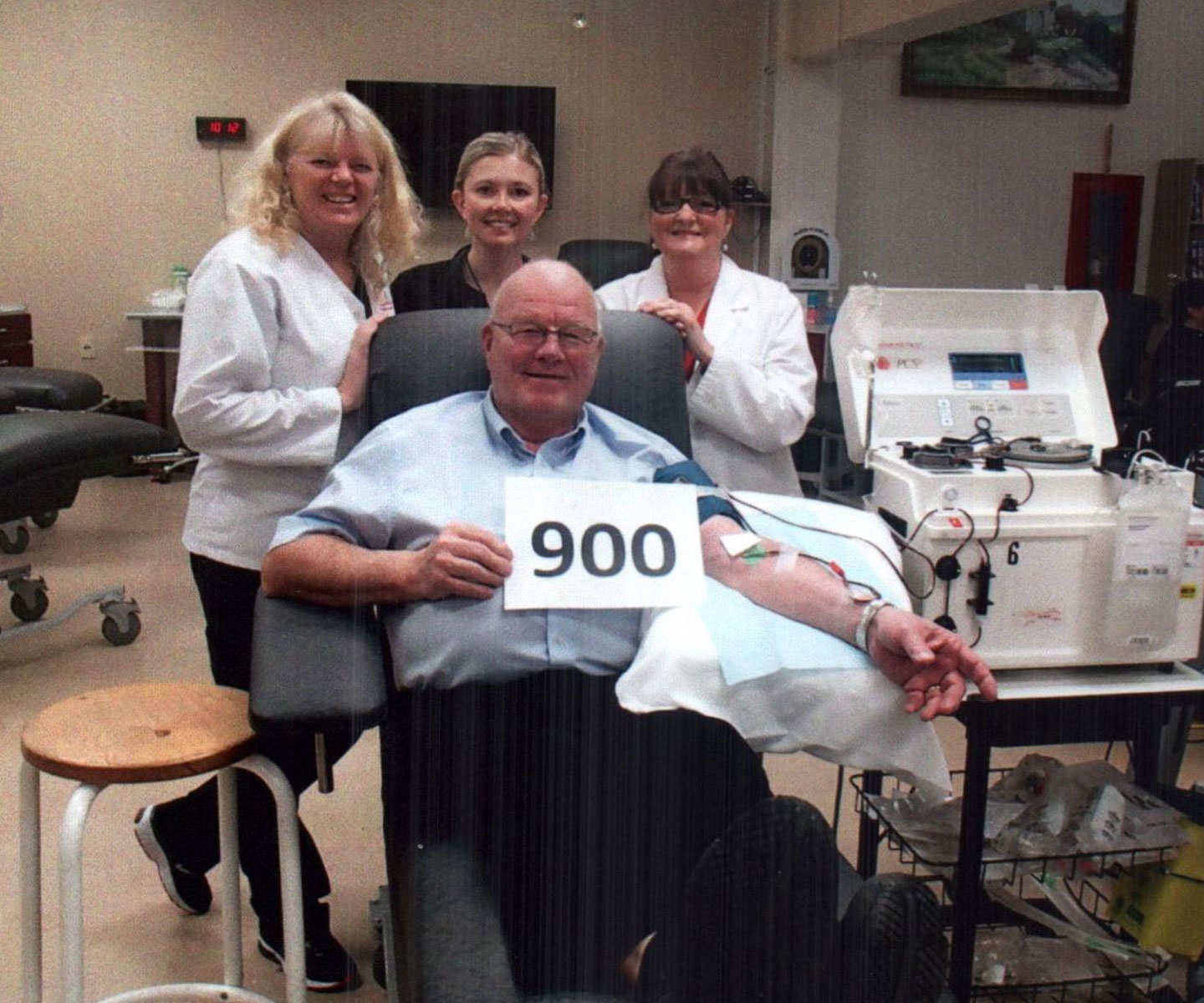 Man sits in blood donor chair holding a sign that says 900, surrounded by staff