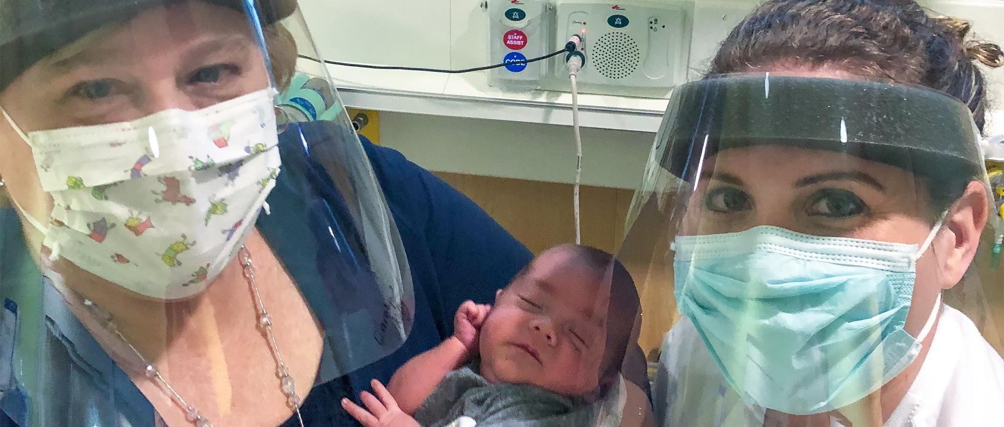 Close up image of Nurse practitioner Carolyn Kelly, Anna Patten and her infant son John-Derrick who required a blood transfusion. Both Carolyn and Anna are wearing masks and shields.