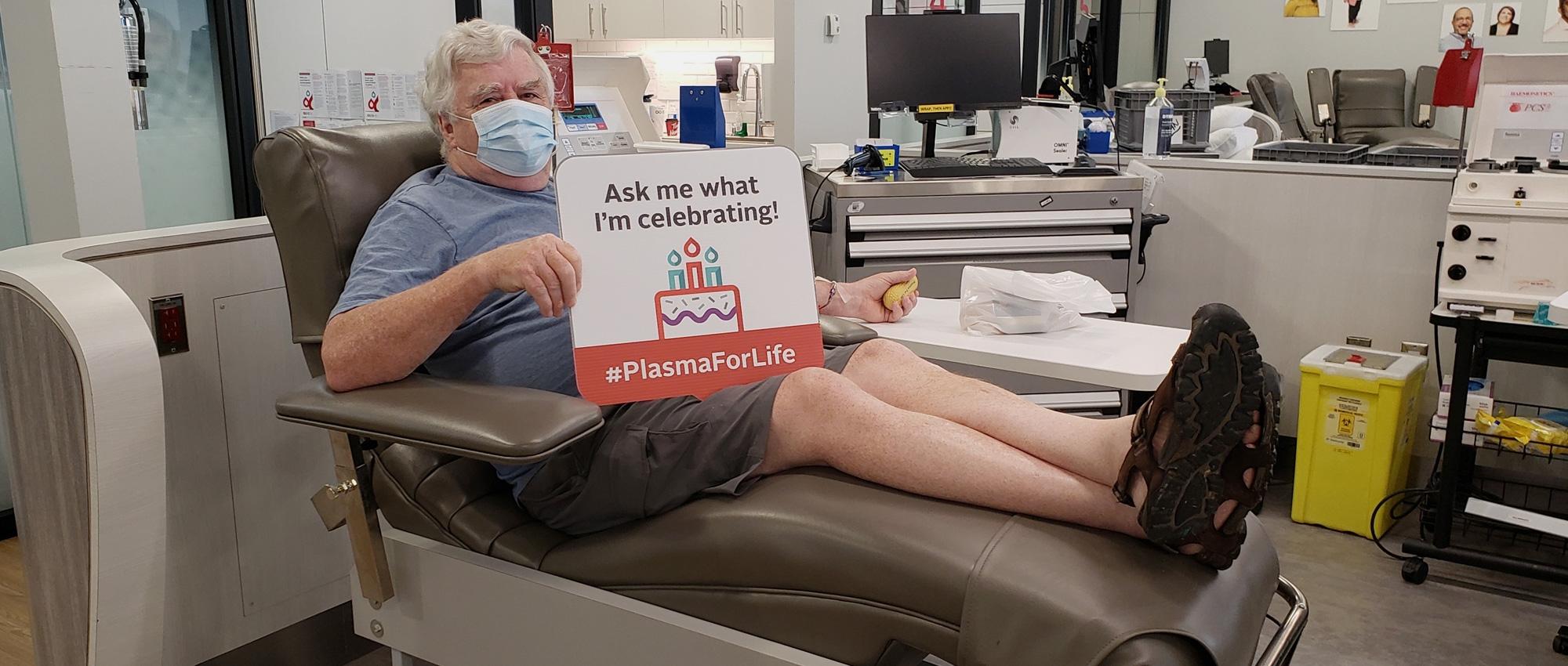 A plasma donor donates plasma while holding a sign that says “Ask me what I’m celebrating!”