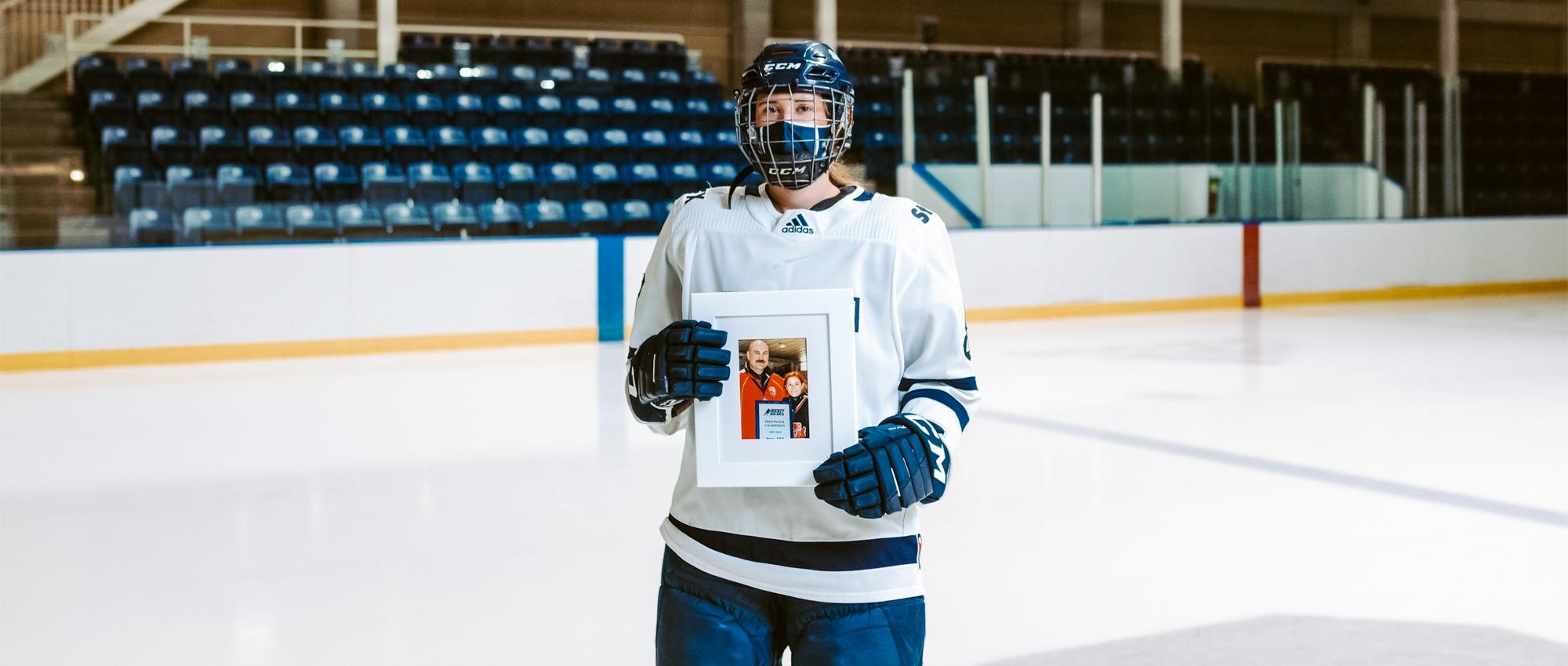 Featured image of Julia Schmitt holding up a picture frame of her and her dad wearing hockey equipment in an indoor hockey rink.