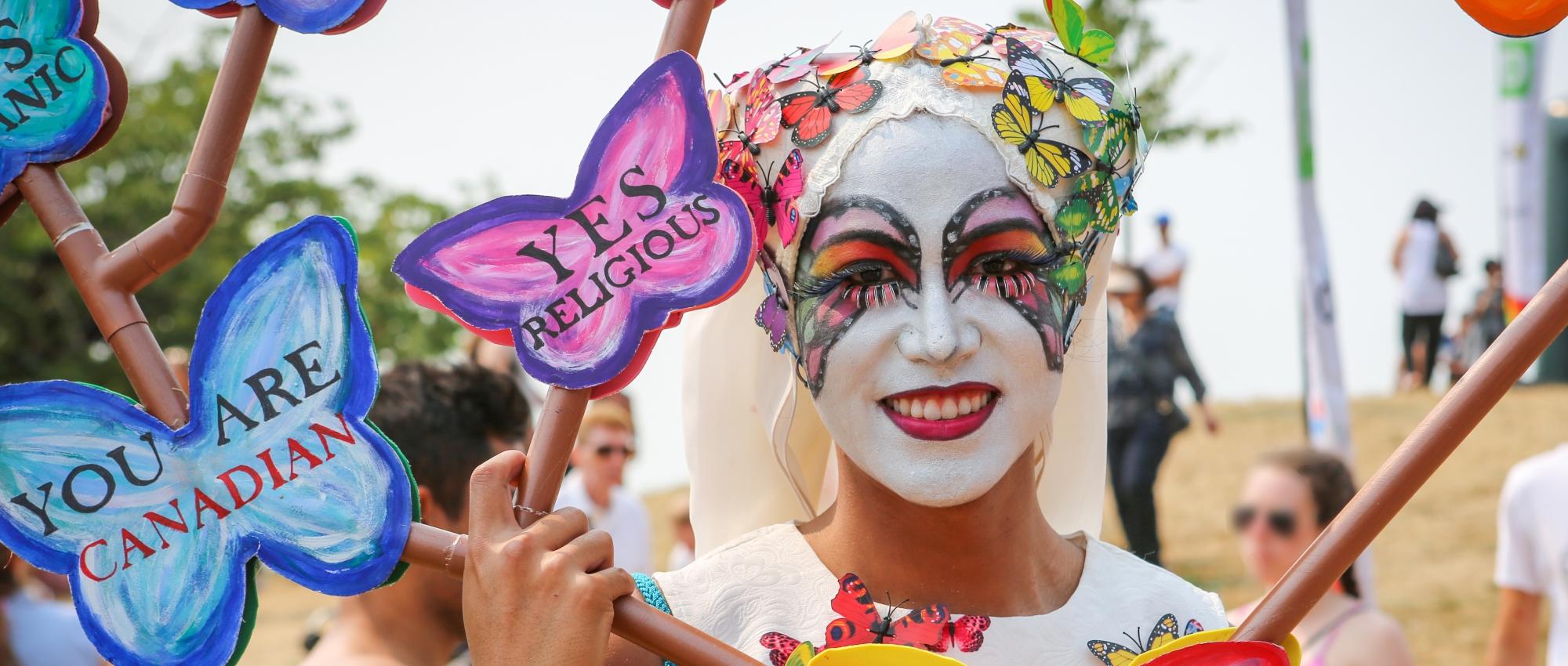 A man in white makeup decorated with colourful butterflies at a beach during a Pride celebration.