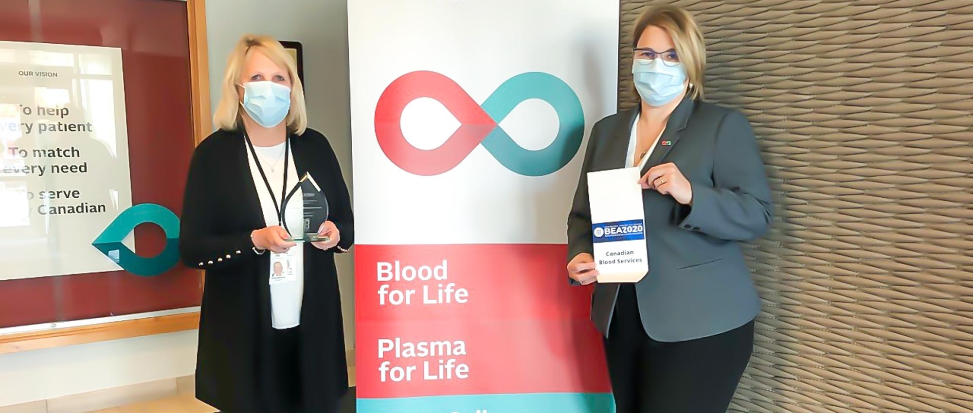 Featured image of director of testing Nancy Angus and director of supply chain operations Andrea Moore holding the award for best organization in Brampton’s life sciences sector standing in front of the Canadian Blood Services pull up stand.