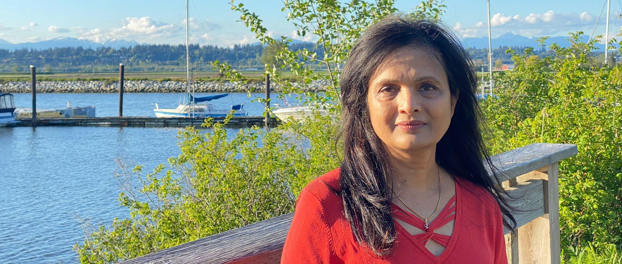 Canadian Blood Services donor care associate in Vancouver’s lower mainland poses on a wooden deck by a lake. There are green plants and boats behind her.