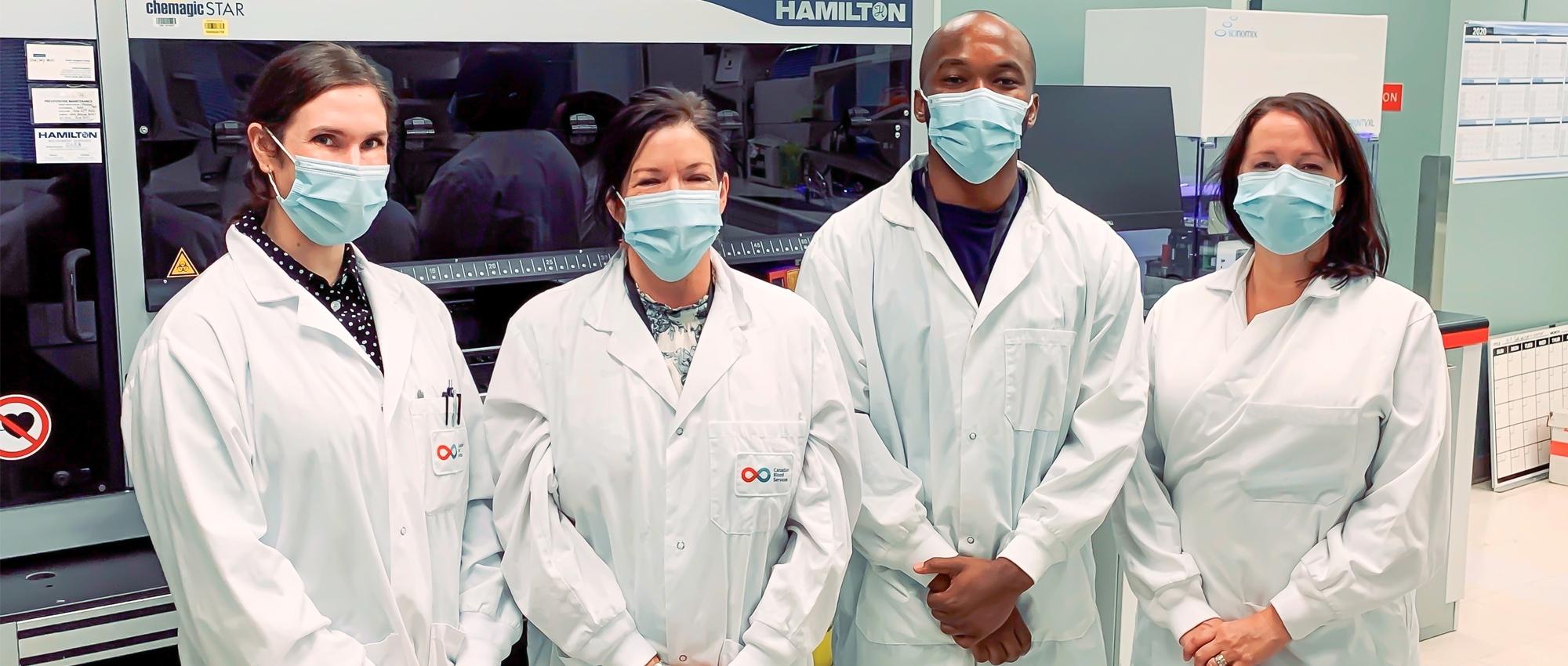 Featured image of lab team members Stacey Vitali, Carissa Kohnen, Andy Tshiula Kalenga and Valerie Conrod standing together in front of a Hamilton machine wearing masks.