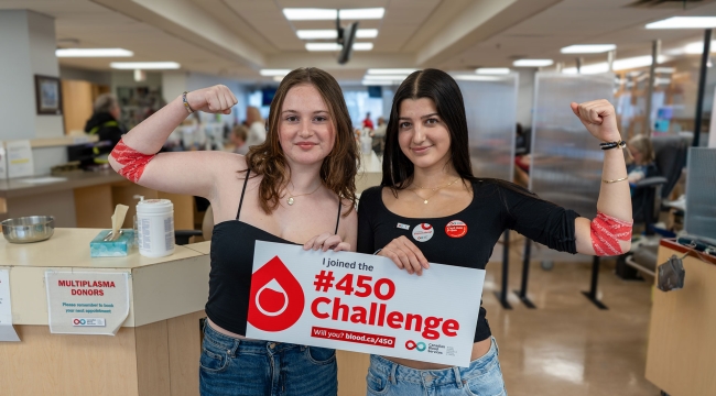 Two first-time blood donors in a donor centre flexing arms with pressure bandages and holding a sign that says “I joined the 450 Challenge”