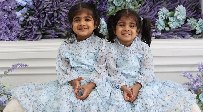 Identical twin girls in blue dresses seated near purple flowers