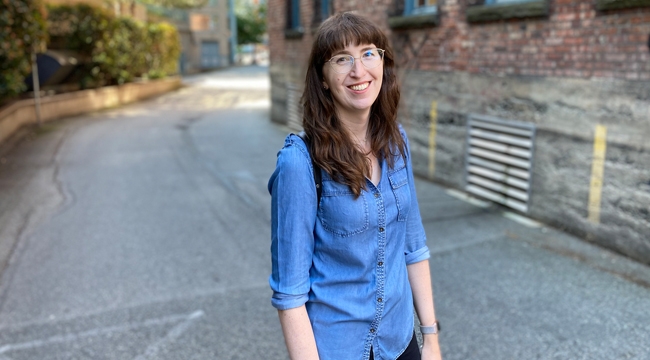 A woman with long brown hair, gold-rimmed glasses and a denim shirt stands on a roadway lined with greenery and brick buildings. 