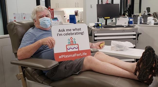 A plasma donor donates plasma while holding a sign that says “Ask me what I’m celebrating!”