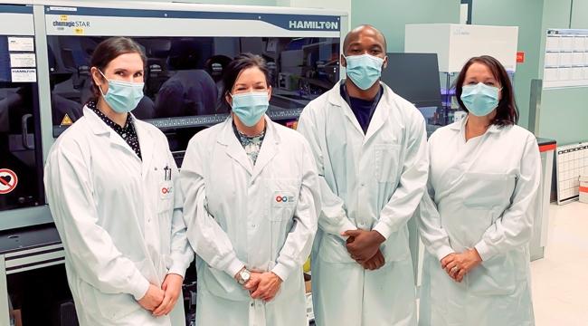 Thumbnail image of lab team members Stacey Vitali, Carissa Kohnen, Andy Tshiula Kalenga and Valerie Conrod standing together in front of a Hamilton machine wearing masks.