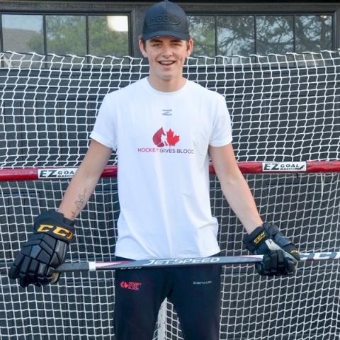 Image of Braden Hache wearing hockey gloves and a Hockey Gives Blood white t-shirt all while holding a hockey stick.