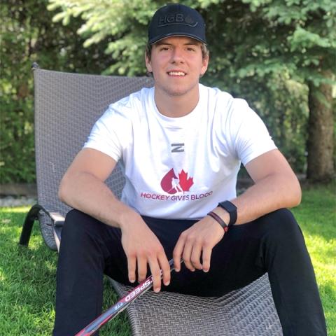 Image of blood donor Cole MacKay sitting outside with black pants and a white Hockey Gives Blood t-shirt on a lawn chair