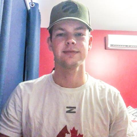Image of blood donor Cole McLaren with a white Hockey Gives Blood t-shirt with a green baseball cap in a bedroom