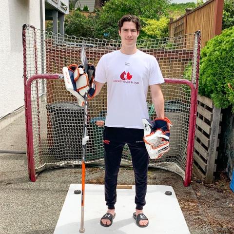 Image of blood donor Dylan Garand wearing hockey gloves and holding a hockey stick standing in front of a goal post outside