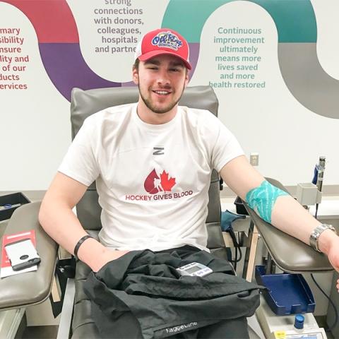 Image of Ethan Cap wearing a red cap and a white t-shirt sitting in a donation chair donating blood at a donor centre