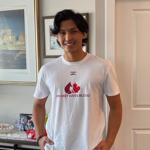 Image of blood donor Alex Kannok Leipert wearing a white hockey gives blood t-shirt with long black hair standing inside the house