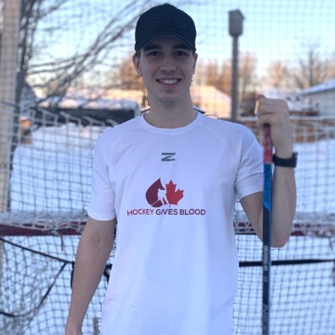 Image of blood donor Tristan De Jong holding a hockey stick standing outside wearing a black cap and white hockey gives blood t-shirt
