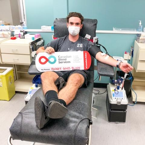 Liam Kindree sitting in a chair donating blood holding a My Reasons sign inside a donor centre wearing a white surgical mask.