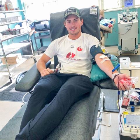 Image of blood donor Logan Nijhoff sitting in a chair donating blood wearing a white Hockey Gives Blood t-shirt.