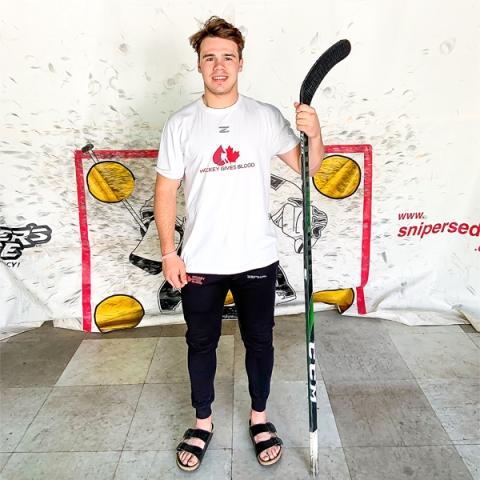 Image of Logan Stankoven with a hockey stick wearing a Hockey Gives Blood T-shirt standing in front of a white tarp with a hockey post.