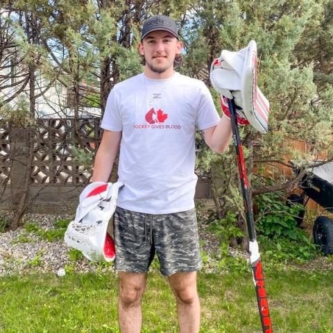 Blood donor Taylor Gauthier holding a hockey stick wearing hockey gloves and a white hockey gives blood t-shirt and camouflage shorts