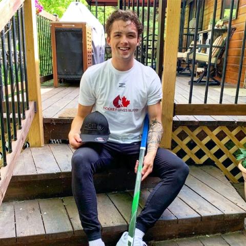 Blood donor Tristen Robins wearing a white t-shirt holding a hockey stick and sitting outside in a backyard patio steps
