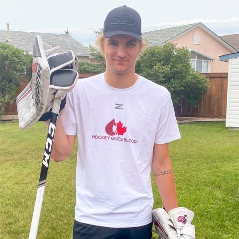 Blood donor Tyler Brennan holding a hockey stick wearing hockey gloves and a white hockey gives blood t-shirt standing outside.
